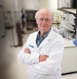 Vern Schramm in his lab, dressed in a white lab coat, standing with his arms folded across his chest.