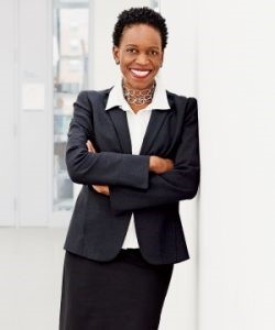 Woman in a black buisness suit with arms crossed standing against a wall and smiling