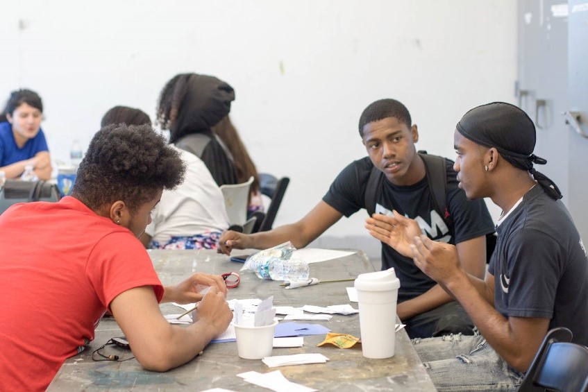 Three male high school students seated at a table with written notes, having a discussion