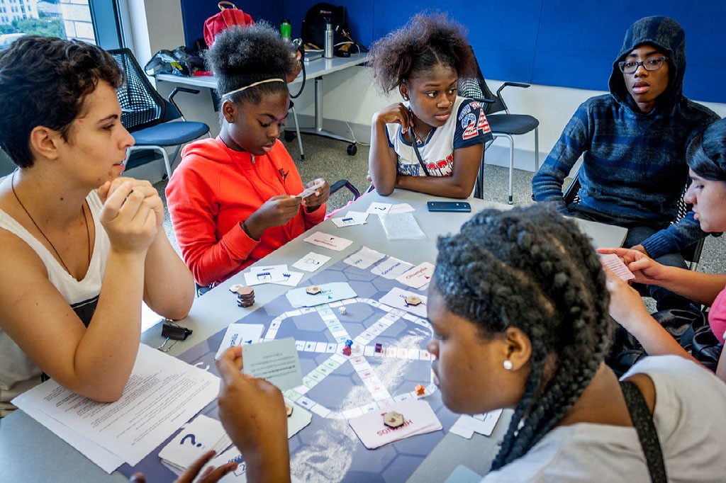 One male and four female high school students seated with a female mentor at a table with a paper test model of a board game sprawled on it. Students are reading from game cards.