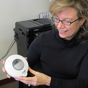 Woman with eyeglasses and black sweater smiling down at a round-shaped, electronic device she's holding