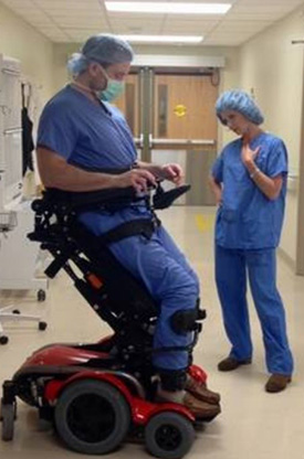 Chris McCulloh on a specialized wheelchair that holds him upright, wearing surgical scrubs and cap, in a hospital hallway alongside Elizabeth Vargas, also in scrubs and cap..