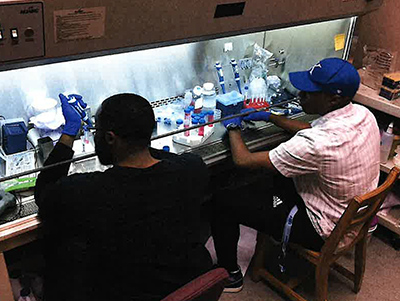 Two male students wearing blue sterilized gloves and seated at a research station, working with vials of red liquid samples.