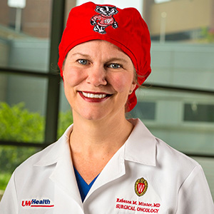 Profile picture of Rebecca Minter wearing a red surgeon's cap with the Bucky Badger mascot and a white surgeon's jacket, smiling.