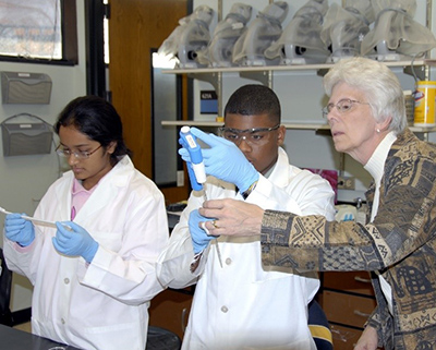 Virginia Shepherd with two students who are wearing lab coats and conducting experiments. She observes the work of one of the students.