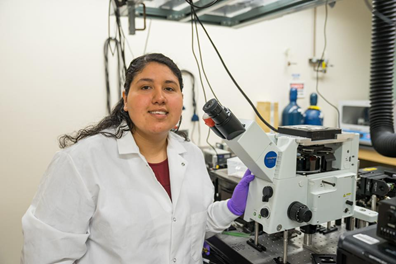 Rosa Romero in a lab, posing in front of a microscope and smiling.