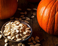 Two large orange pumpkins and a bowl of pumpkin seeds on a wooden table.