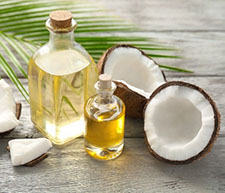 A split-open coconut, two corked glass jars of coconut oil, and a coconut tree leaf on a wooden table.