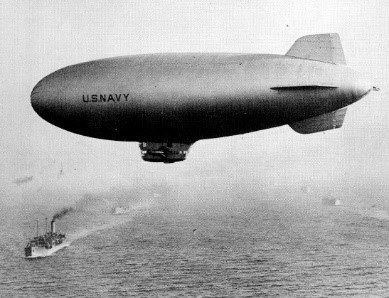A black and white photo of a blimp floating over the ocean and a group of ships.