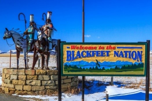 A sign that says “Welcome to the Blackfeet Nation” next to a sculpture of two American Indians on horseback under a blue sky.