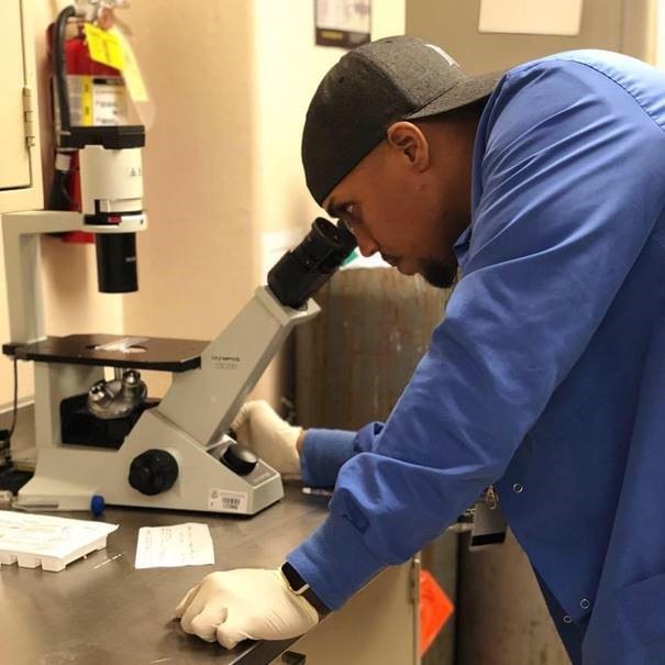Dr. Ramos-Benítez looking into a microscope while wearing a lab coat, gloves, and a baseball cap.
