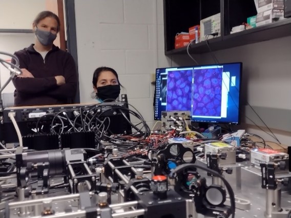 Dr. Munsky standing in the doorway behind a large custom microscope and postdoctoral fellow Dr. Linda Forero Quintero sitting at the computer with images on the screen.