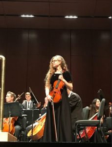 Raven standing onstage with a viola. Other musicians are seated behind her.