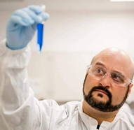 Steve Van Horn wearing a lab coat, gloves, and protective glasses while holding a test tube full of blue liquid.