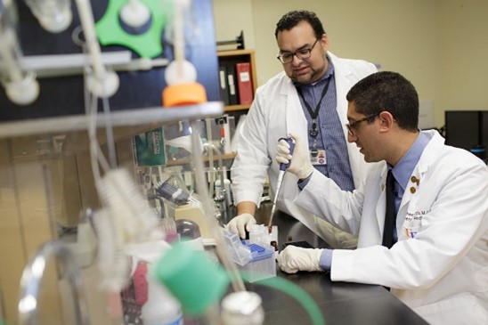 Dr. Guirgis sitting at a lab bench with a pipette while another researcher stands next to him.