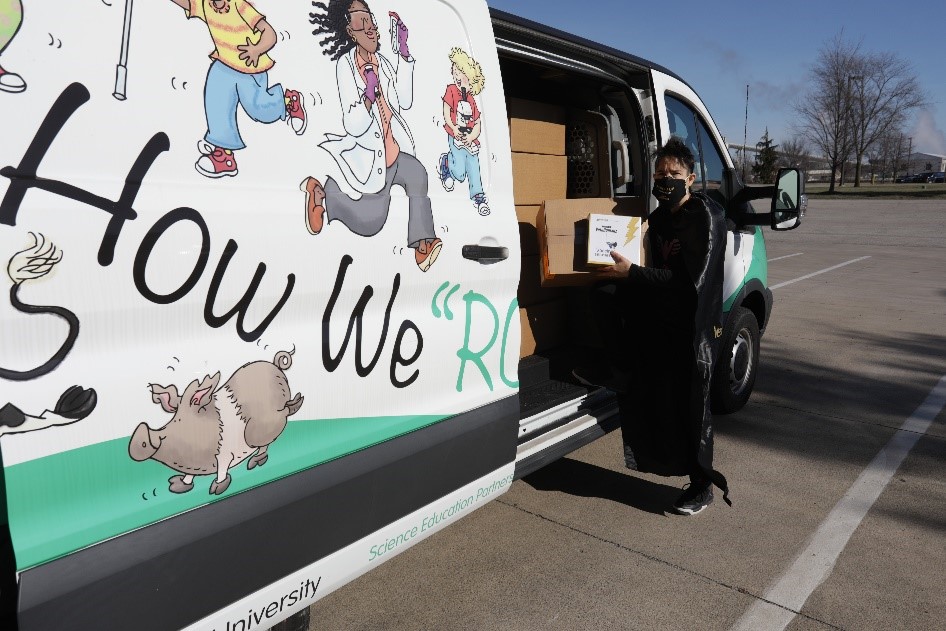 Dr. San Miguel standing in front of a van full of boxes wearing a mask and a cape.