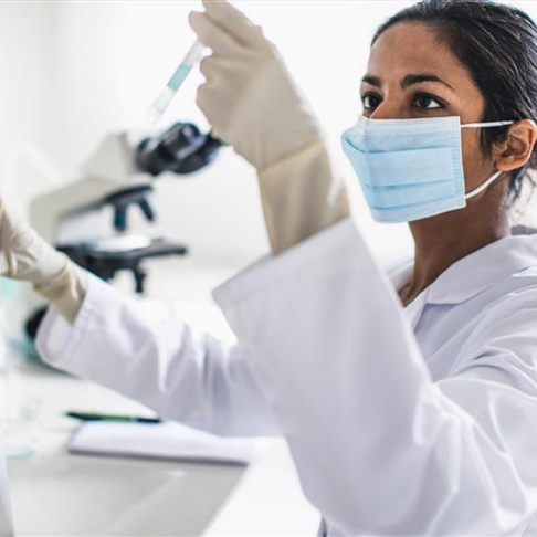 Female scientist wearing a mask and holding a test tube with a microscope in the background.