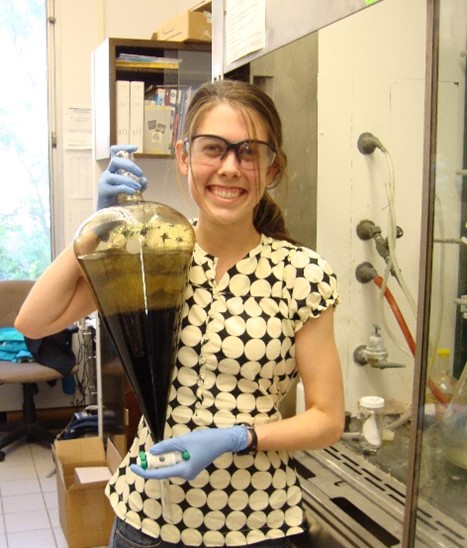 Dr. Parkinson wearing safety glasses and gloves and holding a large glass separatory funnel filled with dark liquid.