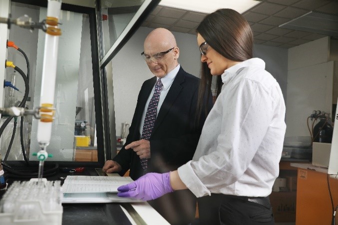 Two people looking at a notebook while standing next to a chemistry fume hood in front of a column that’s collecting liquid in test tubes.