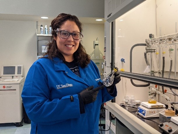 Dr. Andrade standing in front of a chemistry fume hood, holding a glass tube, and wearing a lab coat, gloves, and safety glasses.