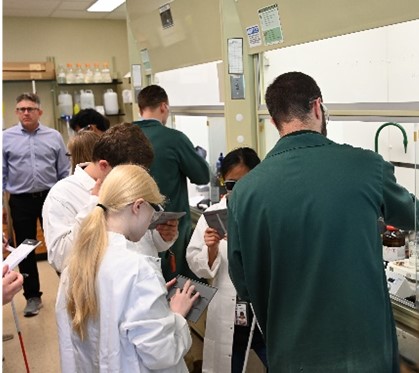 High school students in lab coats and safety goggles feeling tactile graphics while two scientists perform demonstrations of experiments in fume hoods. Dr. Shaw stands in the background.