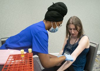 A phlebotomist wearing scrubs, gloves, and a surgical mask drawing blood from Danaé Bartke’s arm.