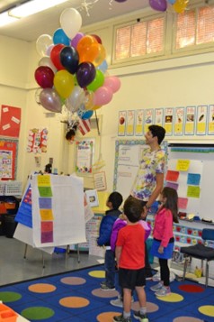 Several kindergarten students and a graduate student watching as a bunch of balloons lift a stuffed animal off the ground.