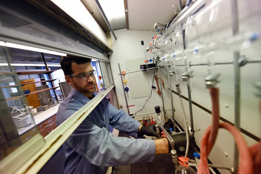 Dr. Leibfarth wearing a lab coat, safety goggles, and nitrile gloves while reaching toward a structure with glass and rubber tubing.