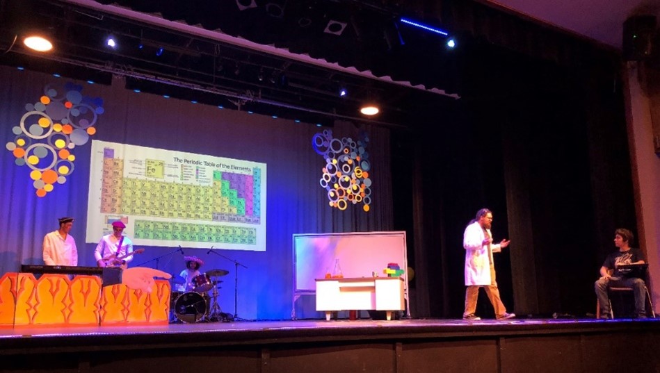 Dr. Queen stands on stage next to a man sitting at a school desk. A band stands nearby. The periodic table of elements is on the wall behind the performers.