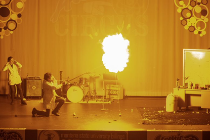 Dr. Queen kneels on stage while holding a long pole that ignites a balloon on fire.
