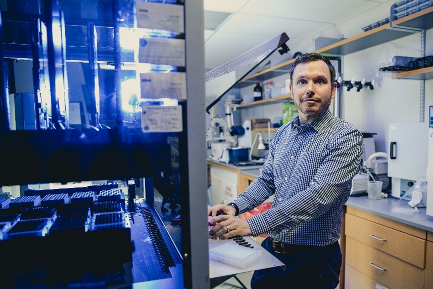 Dr. Gormley looks at the camera while standing at a large piece of lab machinery. Inside, many experiments sit in racks on trays.