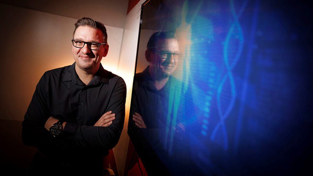 A portrait image of Dr. Helikar standing next to a reflective screen showing a double helix and the outline of a human form.
