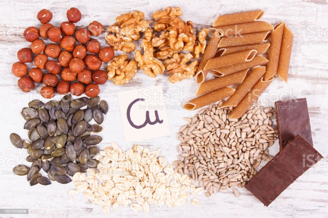 An assortment of sunflower seeds, oats, pine nuts, chocolate, uncooked pasta, walnuts and macadamia nuts surrounding a small piece of paper with copper's symbol Cu written on it.