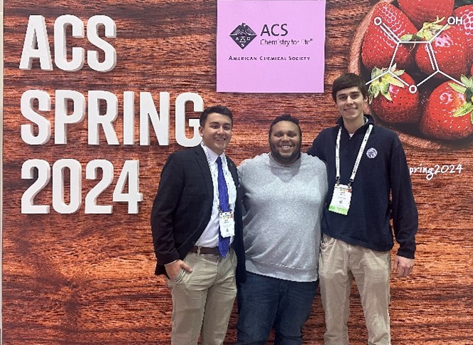 Dr. Gilyot standing side by side with two others in front of a sign that reads ACS Spring 2024.