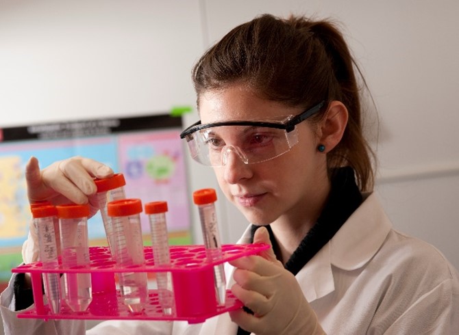 A researcher wearing a lab coat and goggles and looking at test tubes in a rack.