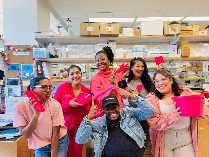 Six people standing in a lab wearing pink clothing and holding various pink items.